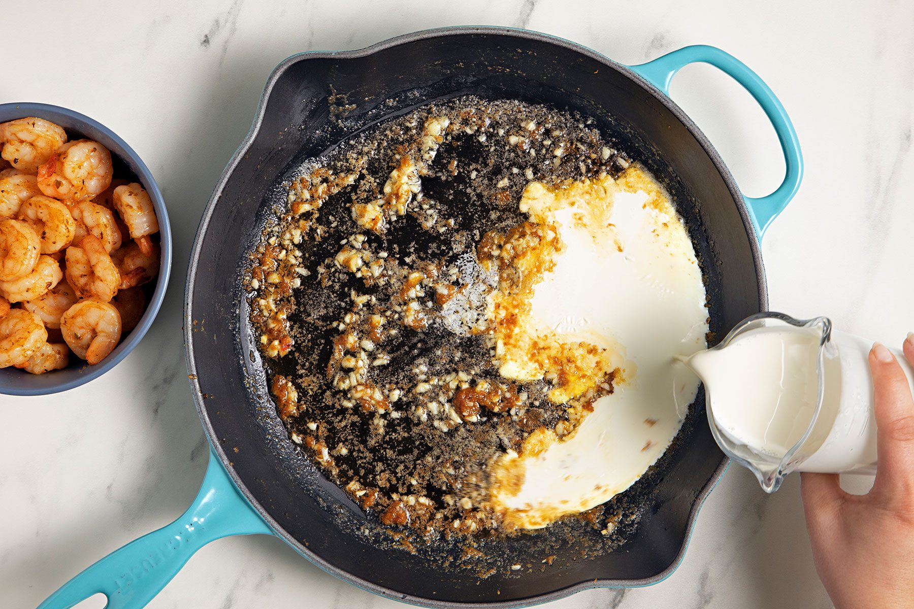 A person is pouring cream from a small pitcher into a black skillet that contains a mixture of spices and ingredients. A bowl of cooked shrimp is placed nearby on the white countertop. The skillet has a teal handle.