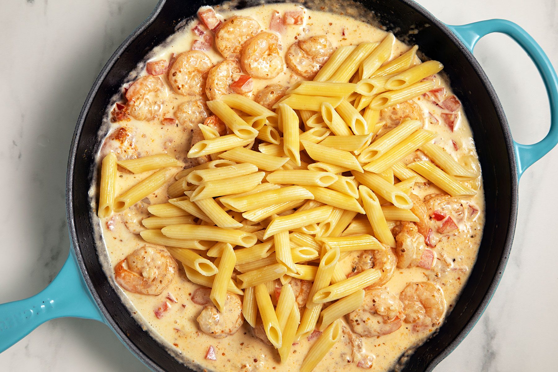 A blue skillet contains a creamy pasta dish with penne, shrimp, and a rich sauce. The sauce is interspersed with chunks of tomatoes and garnished with spices, all on a white countertop.