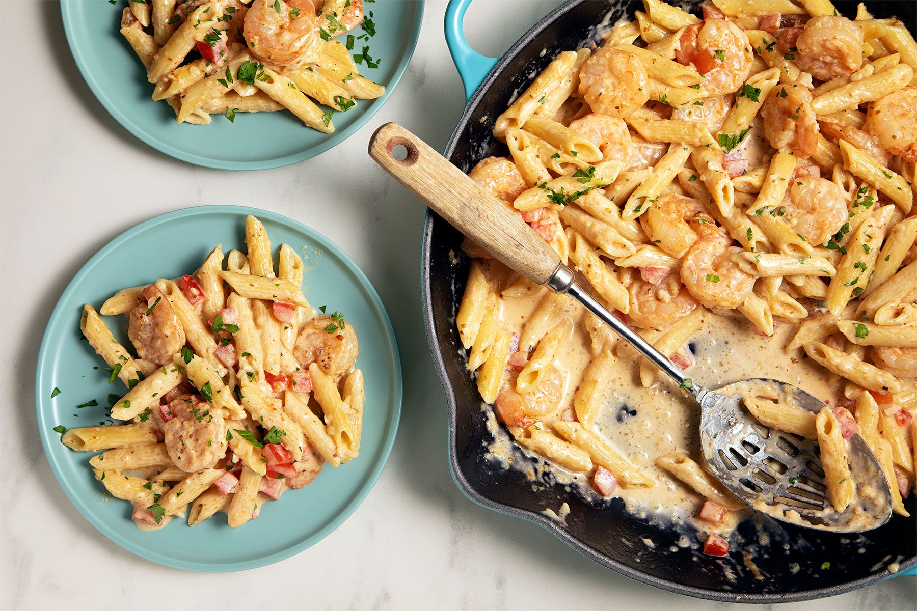 A skillet filled with creamy shrimp pasta sits next to two blue plates, each with a serving of the pasta. The dish includes penne pasta, shrimp, and chopped tomatoes, garnished with fresh herbs. A slotted spoon rests in the skillet.