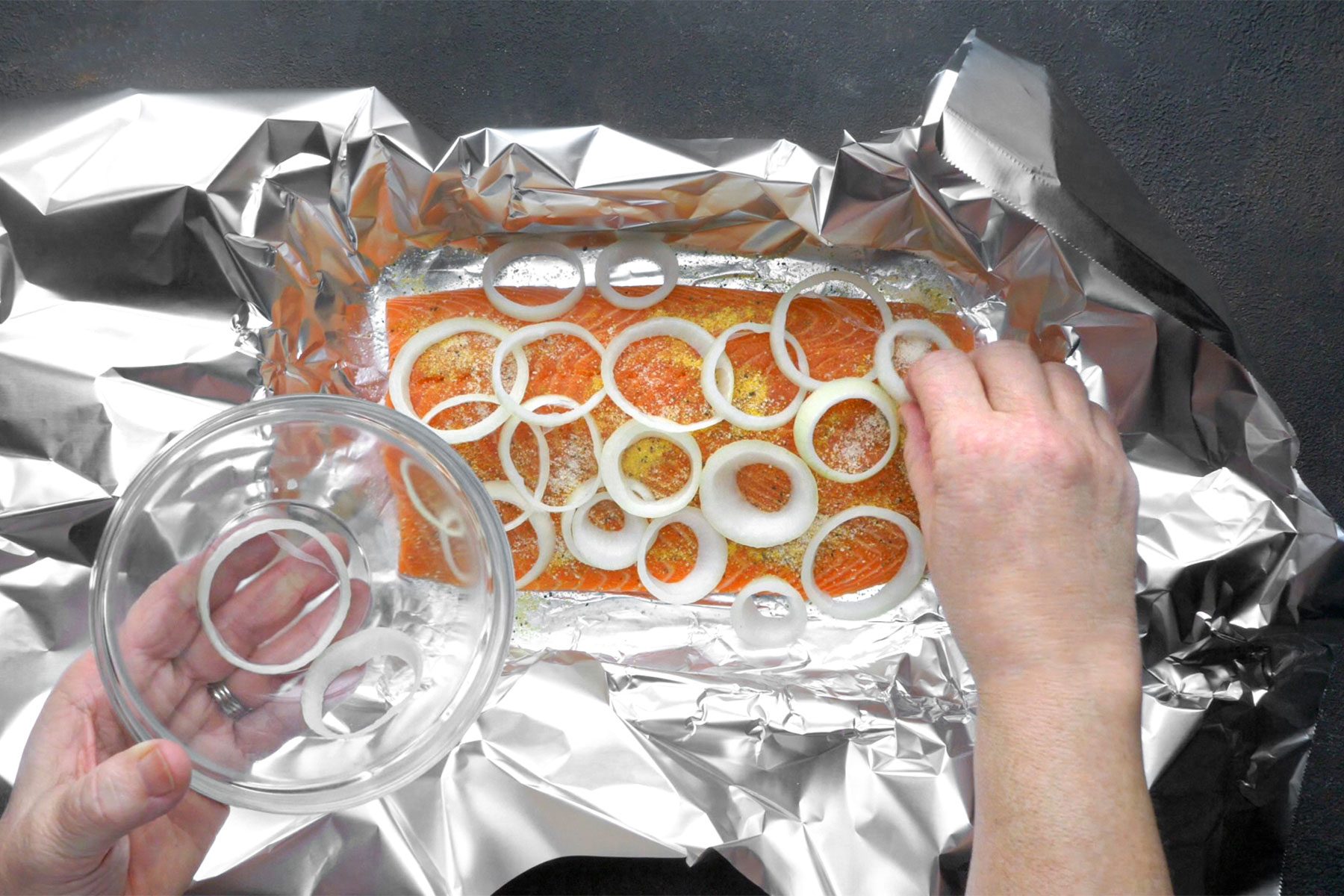 Overhead shot of baking pan with heavy-duty foil; grease lightly; placed salmon skin side down on foil; sprinkled with lemon pepper and onion salt; top with onion and lemon; dark grey background;