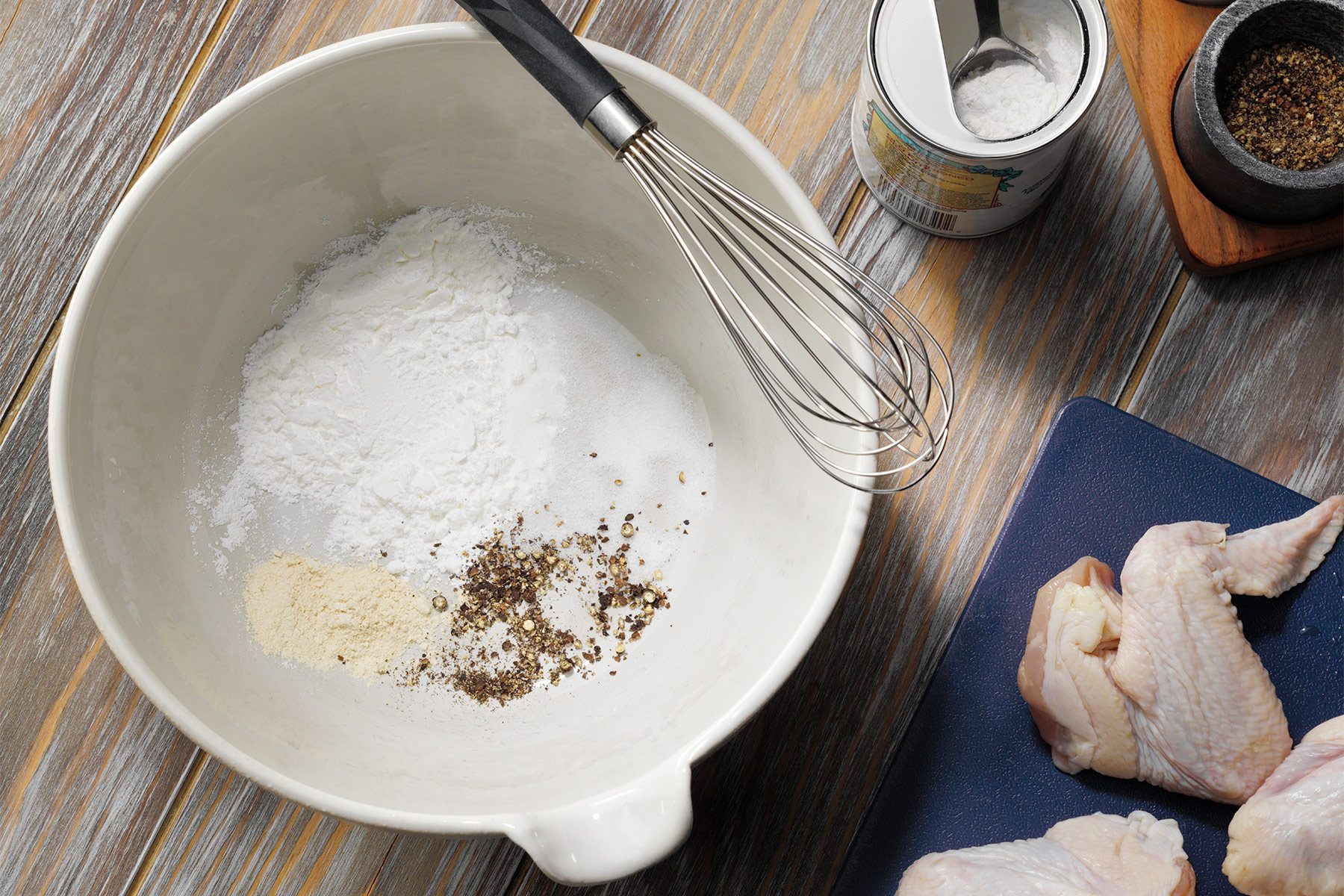 A white mixing bowl filled with flour and spices, including black pepper and possibly garlic powder, is on a wooden surface. Beside it is a whisk, an open can of baking powder with a measuring spoon, and a cutting board with pieces of raw chicken.