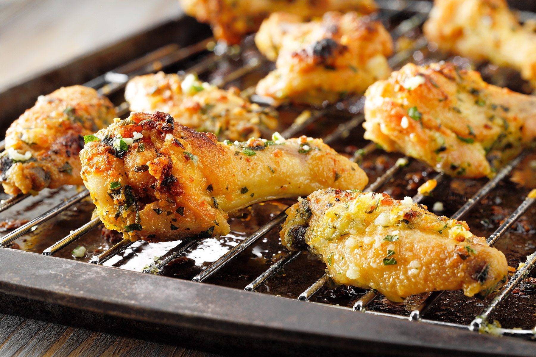 Grilled chicken wings coated with herbs and spices are arranged on a rack over a baking sheet. The wings are golden brown and crispy, showing char marks, with small bits of chopped green herbs visible on the surface.
