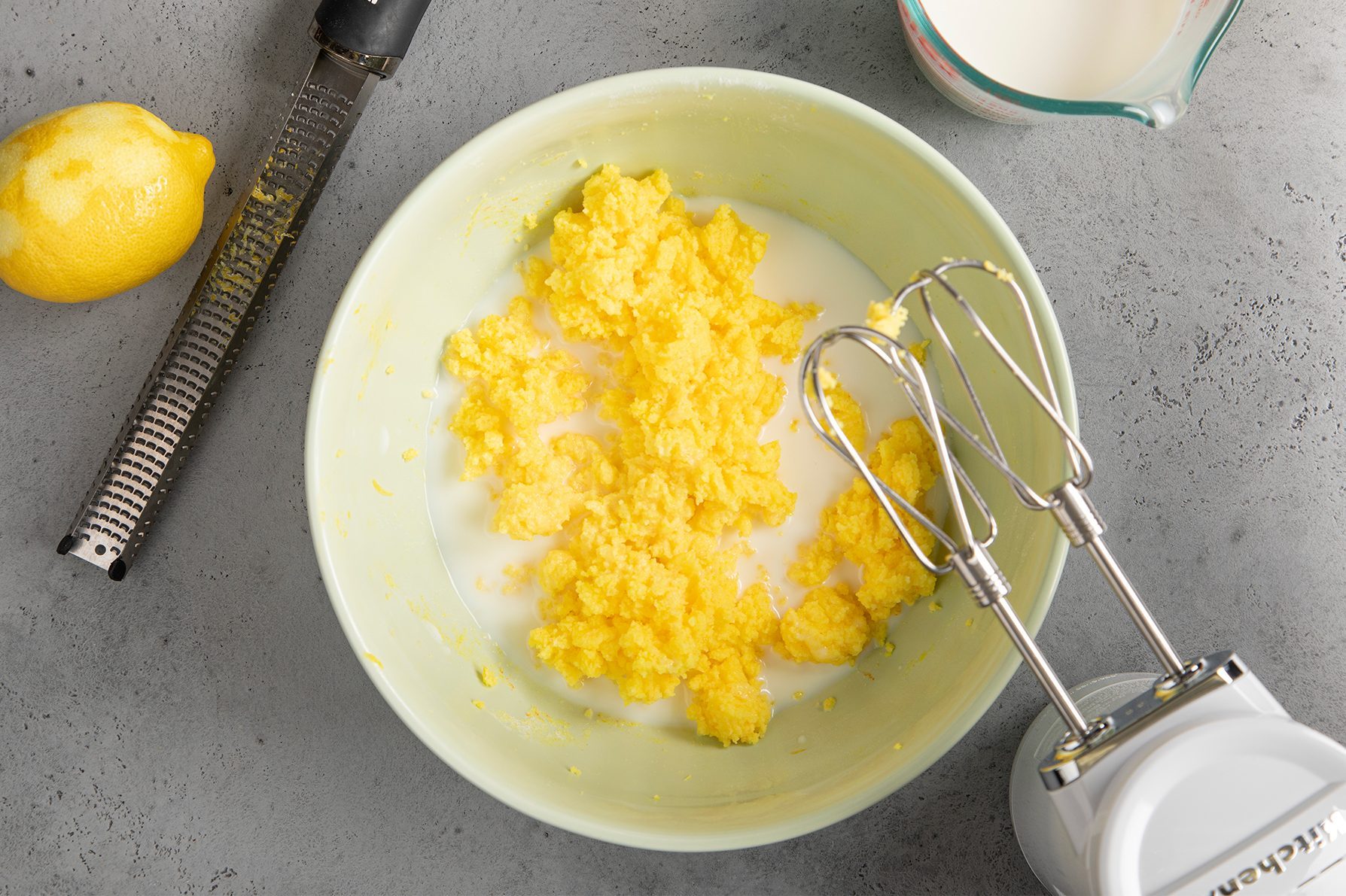 A mixing bowl filled with a bright yellow, coarse mixture sits on a gray countertop. Beside it are a lemon, a grater, a measuring cup with liquid, and a hand mixer with beaters partially submerged in the bowl's contents.