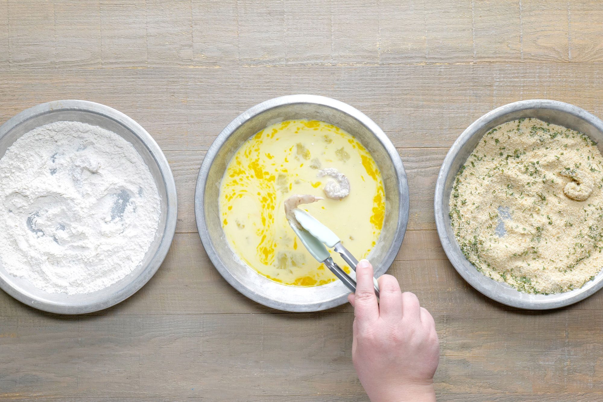 Overhead shot of whisk eggs and milk in a separate shallow bowl; mix flour salt and pepper in a third shallow bowl; dip shrimp in flour to coat both sides; shake off excess; dip in egg mixture; then in bread crumbs; tongs; wooden background;