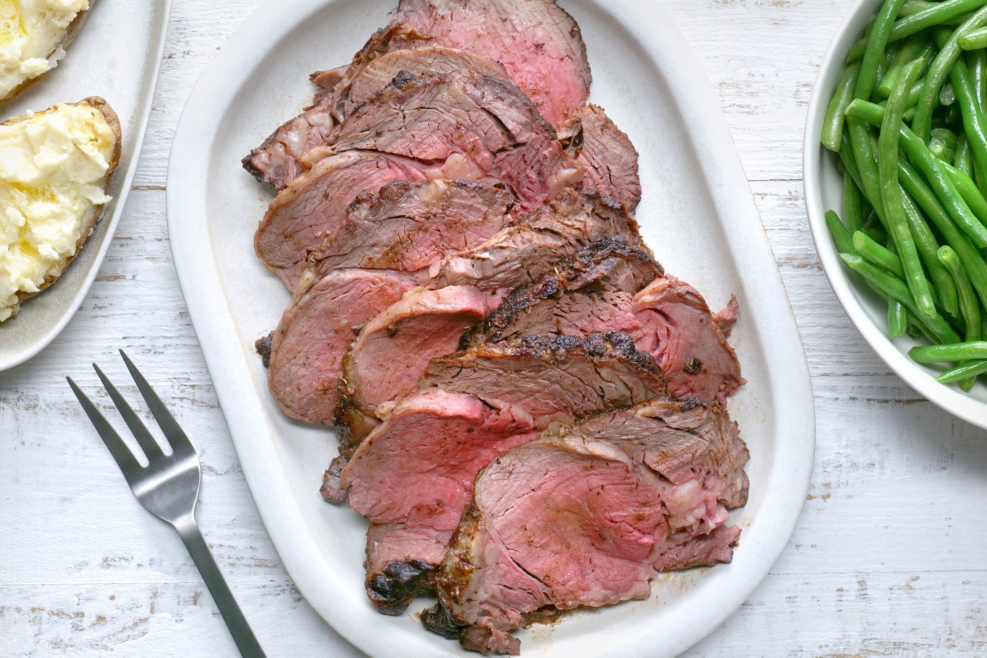 Top view shot of Seasoned Ribeye Roast; serve on large wooden platter; with fresh beans and mashed potatoes; fork; white wooden background;
