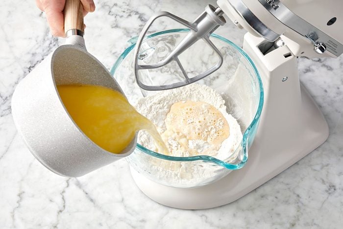 wide shot of pouring yellow mixture into a glass bowl; stand mixer; marble surface;