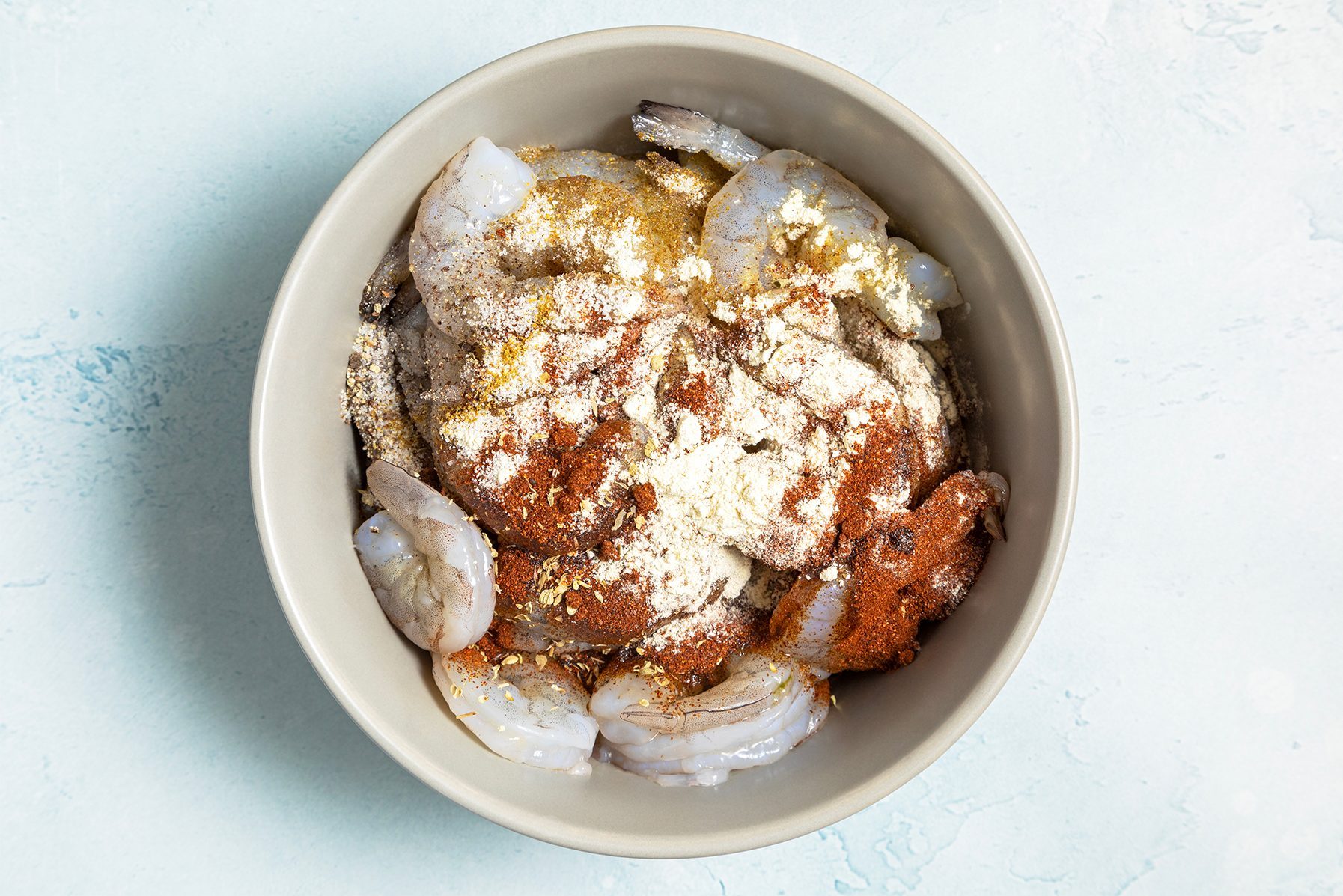 A bowl of raw shrimp seasoned with various spices, including paprika, garlic powder, and possibly other herbs and seasonings.