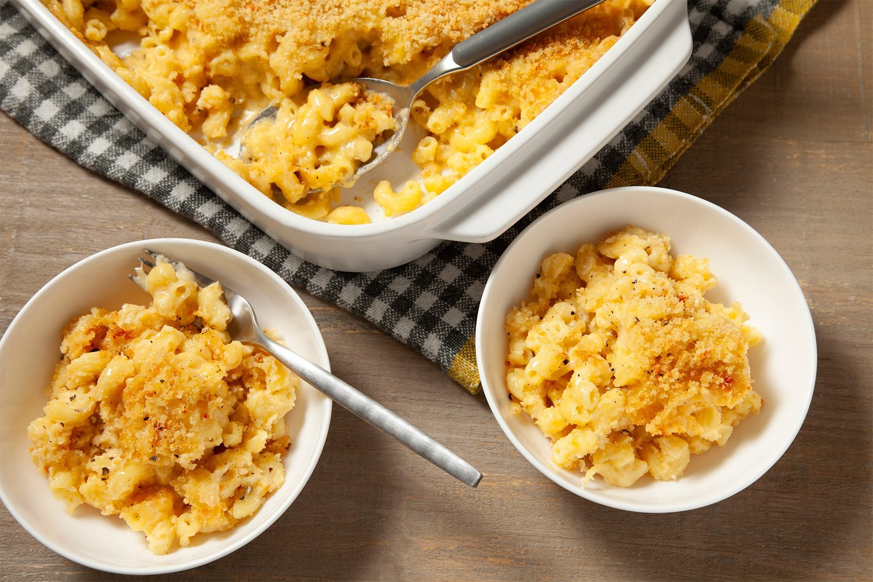 3/4th shot; wooden background; Baked Three-Cheese Macaroni served in two small white bowls with fork; Baked Three-Cheese Macaroni in rectangular dish over kitchen napkin with spoon inside it;
