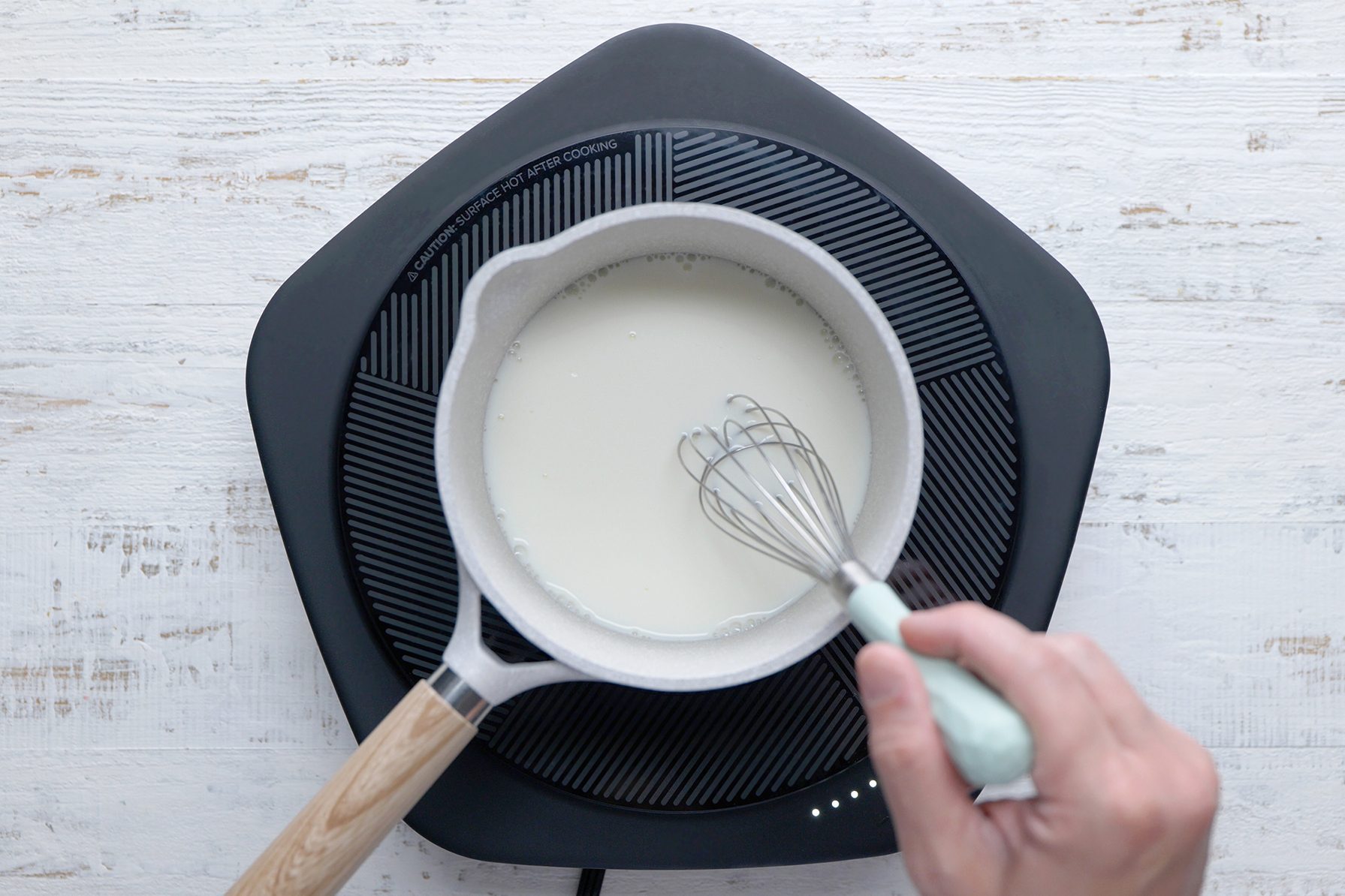 A person's hand holding a light teal whisk is stirring a white sauce in a white saucepan placed on a black induction cooktop. 