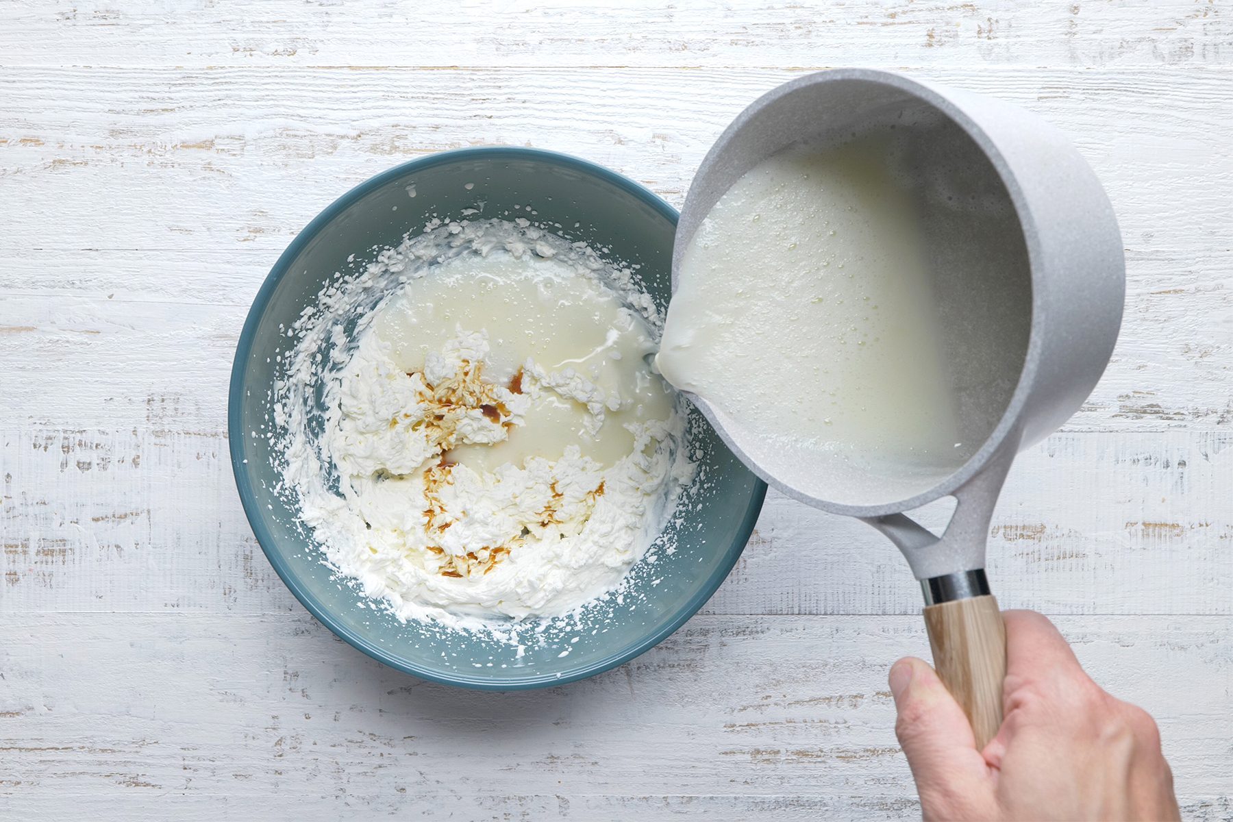 A person is pouring liquid from a saucepan into a mixing bowl containing flour and possibly other ingredients, on a white wooden surface.