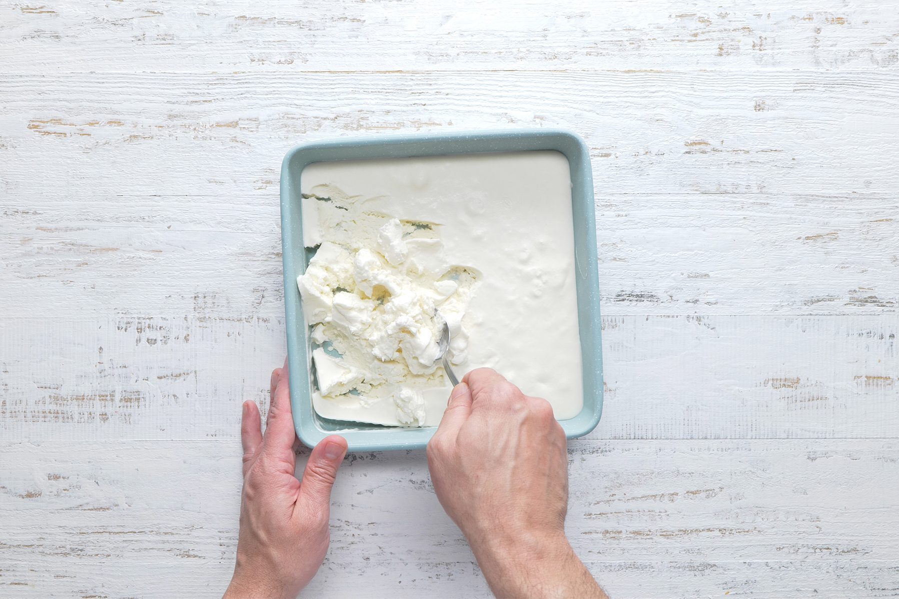 A person’s hands are seen holding a spoon and cutting into a square pan filled with a creamy white mixture, likely a dessert or dairy product, positioned on a white wooden surface.