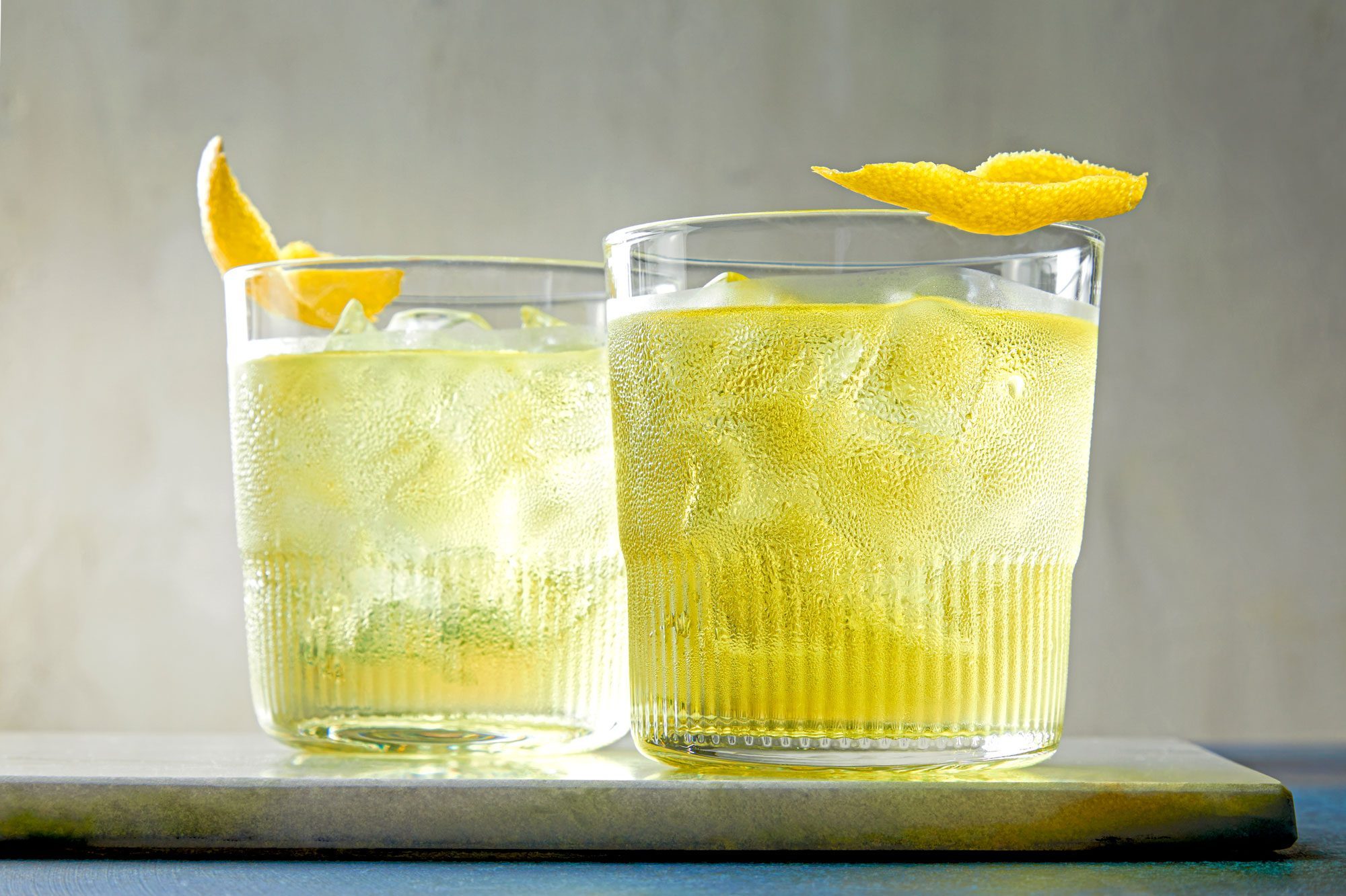 Low angle view shot of White Negroni; served in two glasses; garnished with lemon peel twist; on wooden tray; grey background;