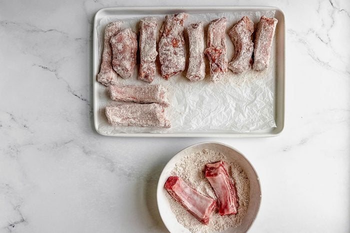 Floured ribs for Taste of Home fried ribs on a white tray and in a white bowl on a marble surface.