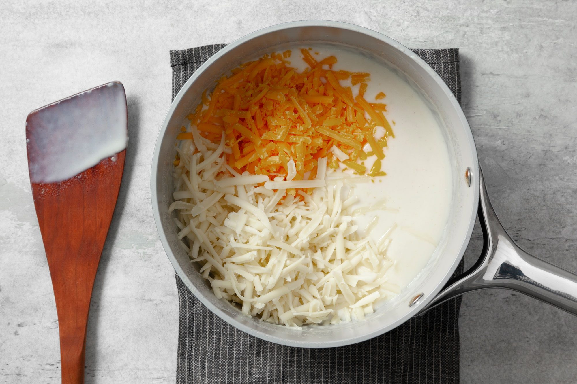 Overhead shot of stir in 1 cup cheddar cheese; and swiss cheese until melted; wooden spatula; kitchen cloth; grey marble background;