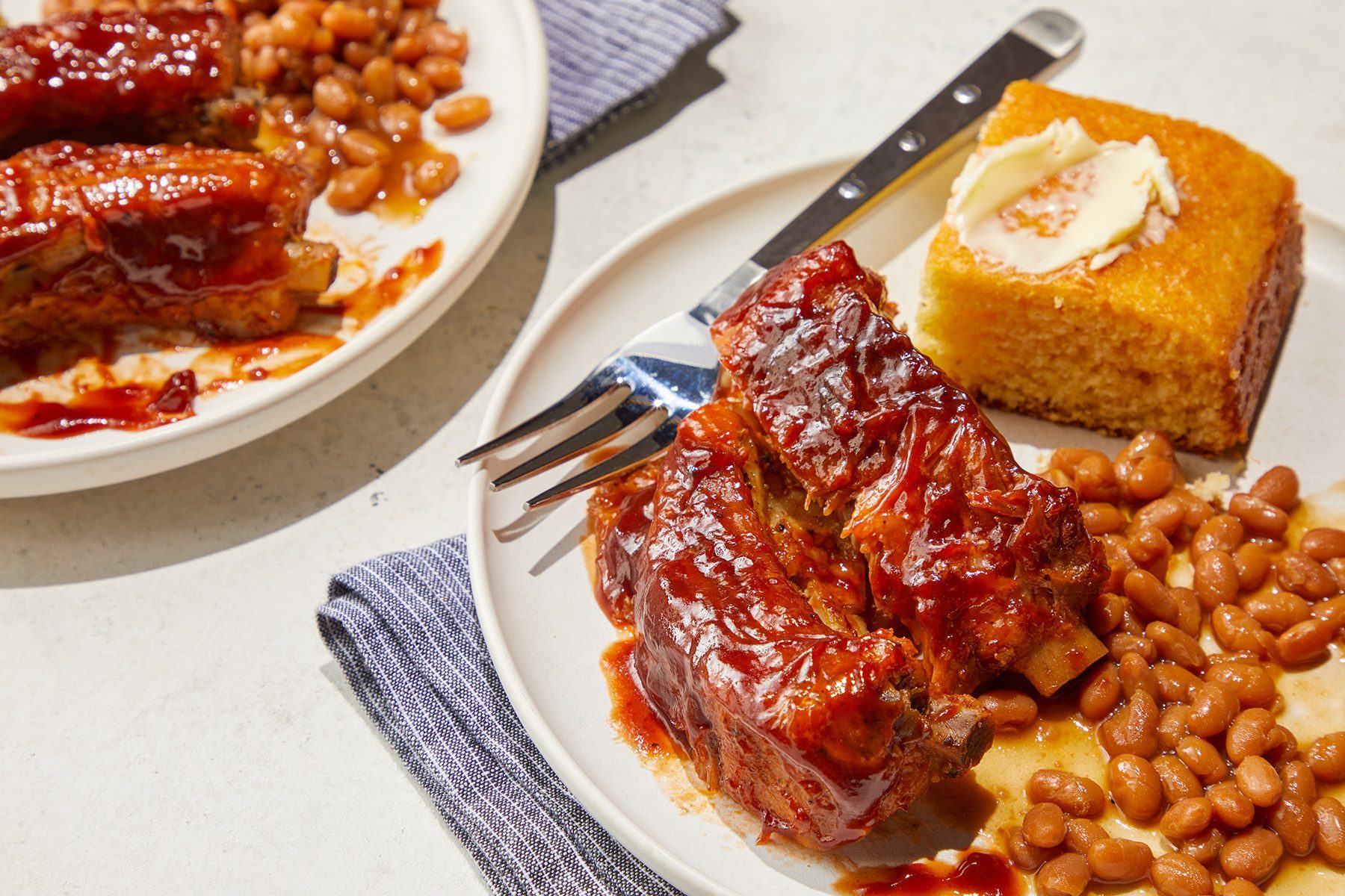 High angle view shot of Slow-Cooker Baby Back Ribs; served on plates; with beans; cornbread and muffin; fork; napkin; marble background