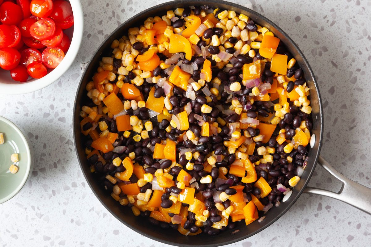 onion, garlic, black beans, corn and orange pepper in a saute pan with a bowl of cherry tomatoes on the surface to be added