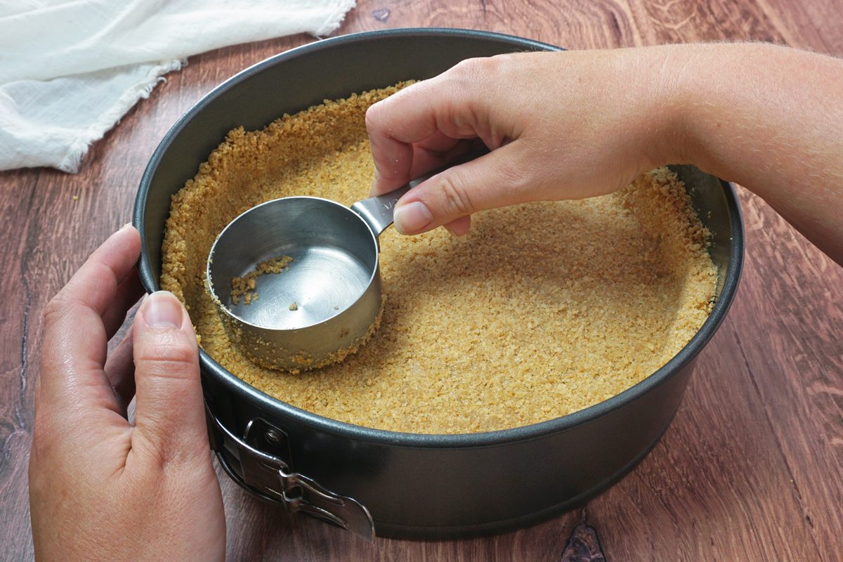 Using a measuring cup to press a graham cracker crust into a springform pan to make amaretto cheesecake RMS 91184 for Taste of Home