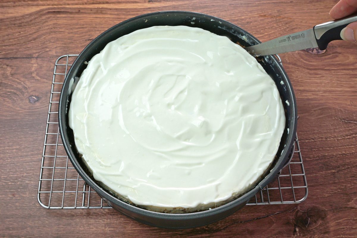 An amaretto cheesecake cooling on a wire rack while a knife loosens the side for Taste of Home RMS 91184