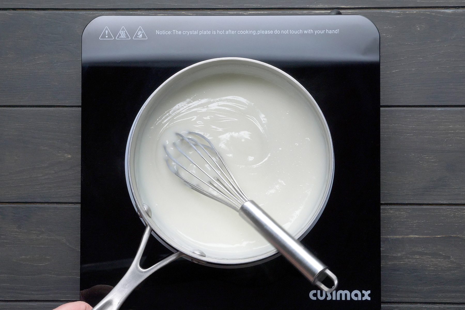 A saucepan sits on an induction stove, containing a white liquid being stirred with a whisk. The induction stove has the brand name "Cusimax" on it and a warning message indicating that the crystal plate is hot after cooking and should not be touched by hand.