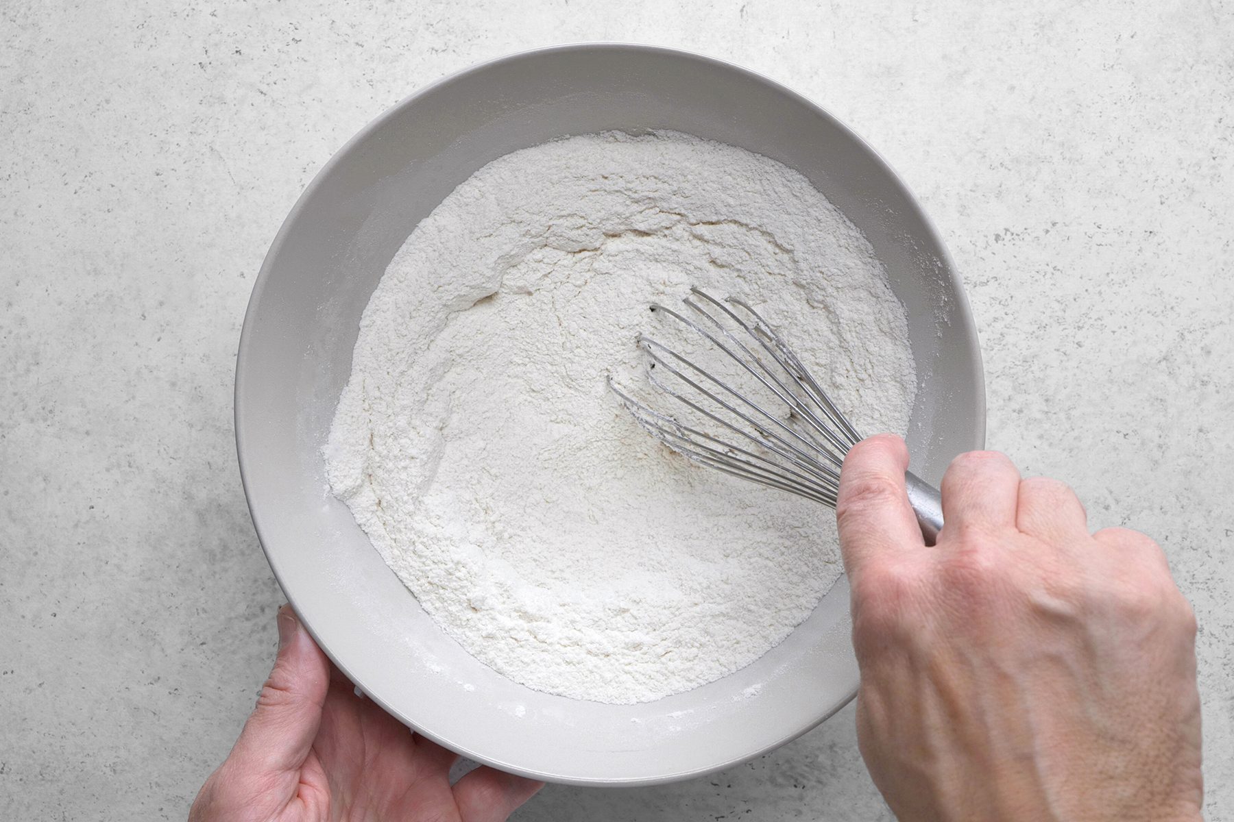 A person's hand is whisking flour in a large, light gray bowl. The bowl sits on a speckled, light gray surface. The flour is fluffy and finely sifted.