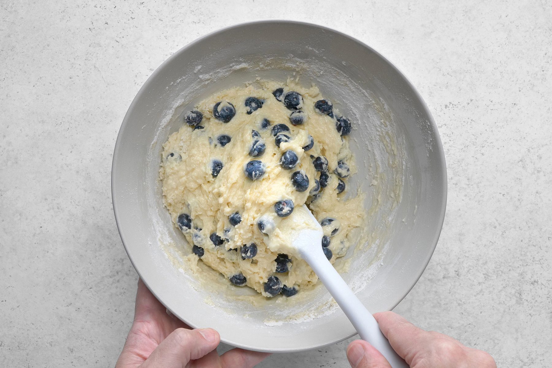 A bowl containing a thick batter mixed with blueberries is being stirred with a white spatula. The bowl is being held by two hands over a light gray countertop.