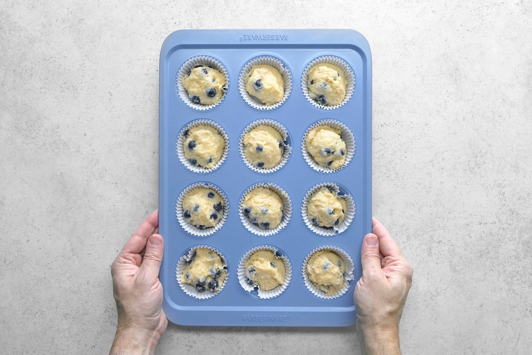 Two hands holding a blue muffin tray with twelve compartments filled with blueberry muffin batter in white paper liners, ready to be baked. The background is a light, textured surface.