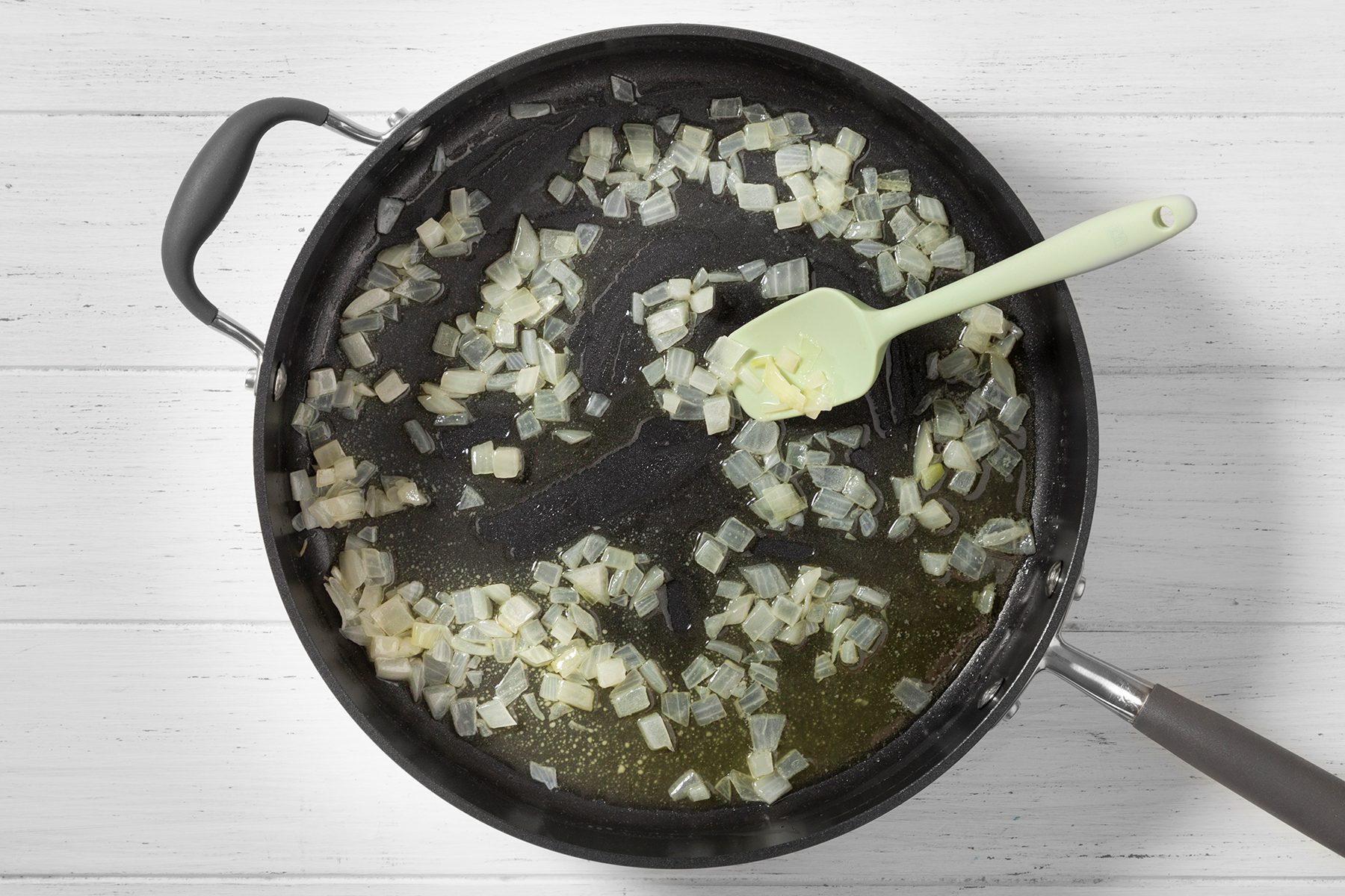 A black skillet on a white wooden surface containing sautéed onions being stirred with a light green spatula. The onions are softened and translucent, with a small amount of liquid visible in the pan.