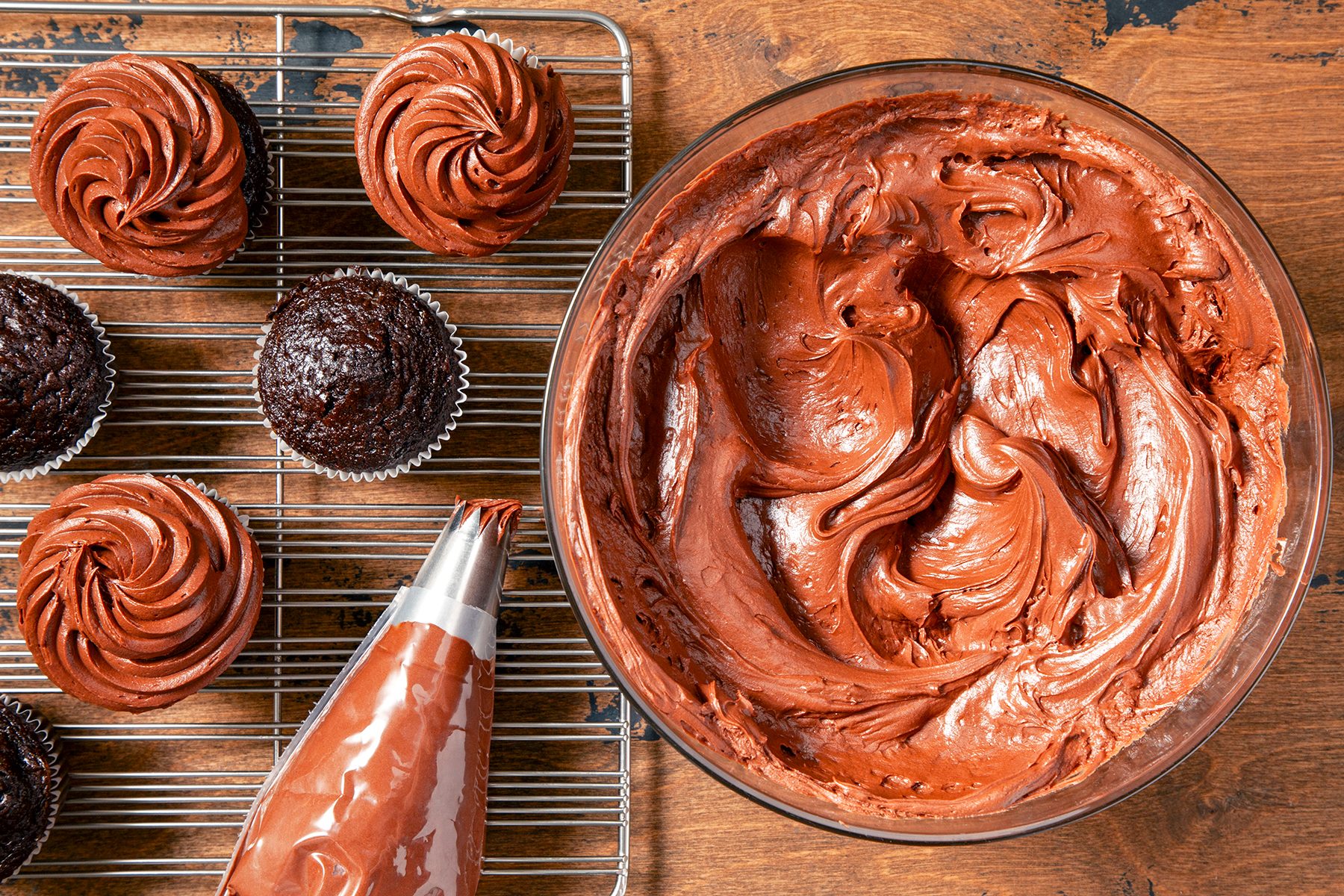 A large bowl filled with smooth, chocolate frosting sits on a wooden surface. Nearby, a wire cooling rack holds several chocolate cupcakes, some of which are already frosted. A piping bag filled with chocolate frosting lies beside the bowl and the cupcakes.