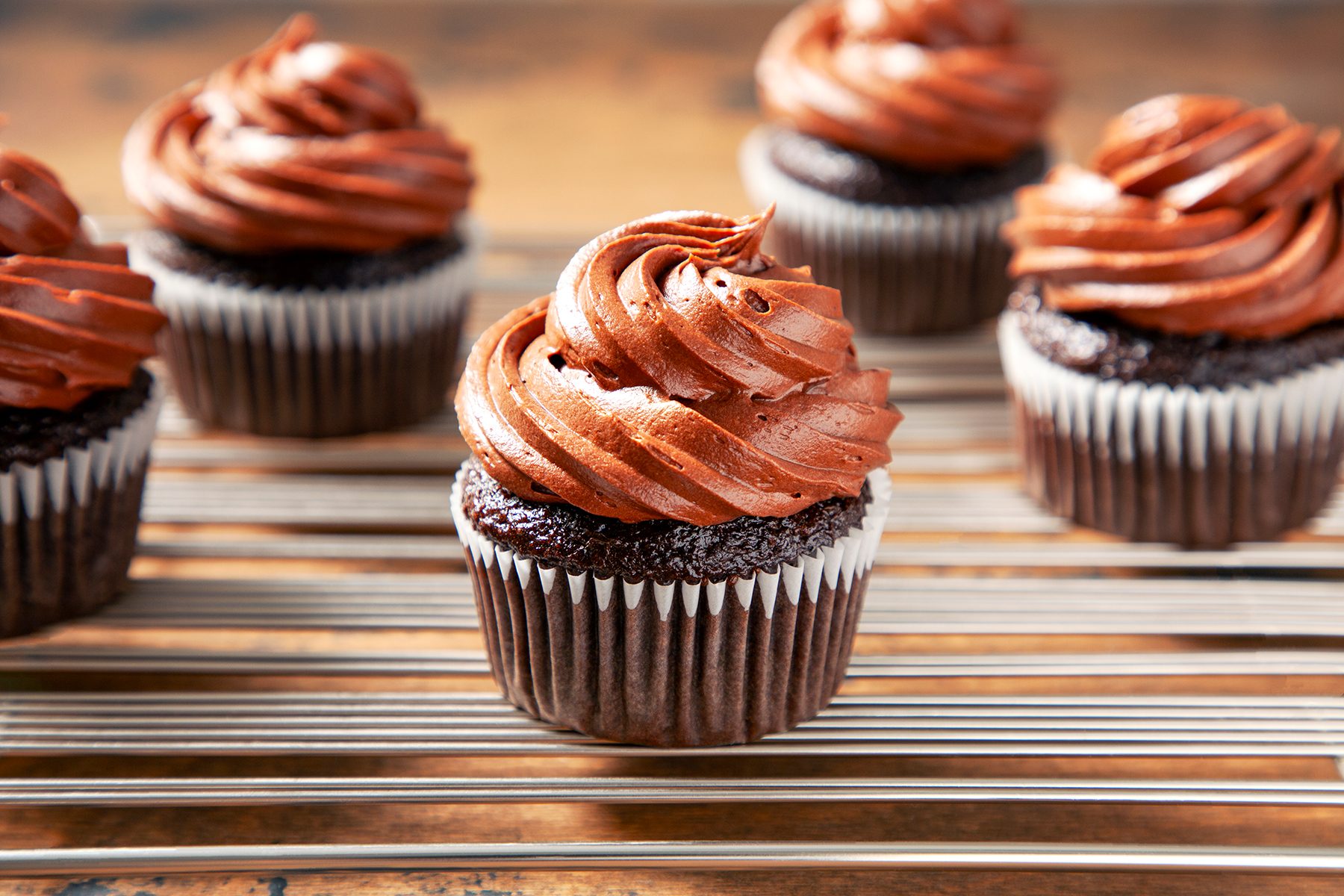 Close-up of five chocolate cupcakes topped with swirls of chocolate frosting, placed on a cooling rack. The cupcakes are displayed in a well-lit setting, emphasizing the rich texture of the frosting and the moist appearance of the cupcakes.