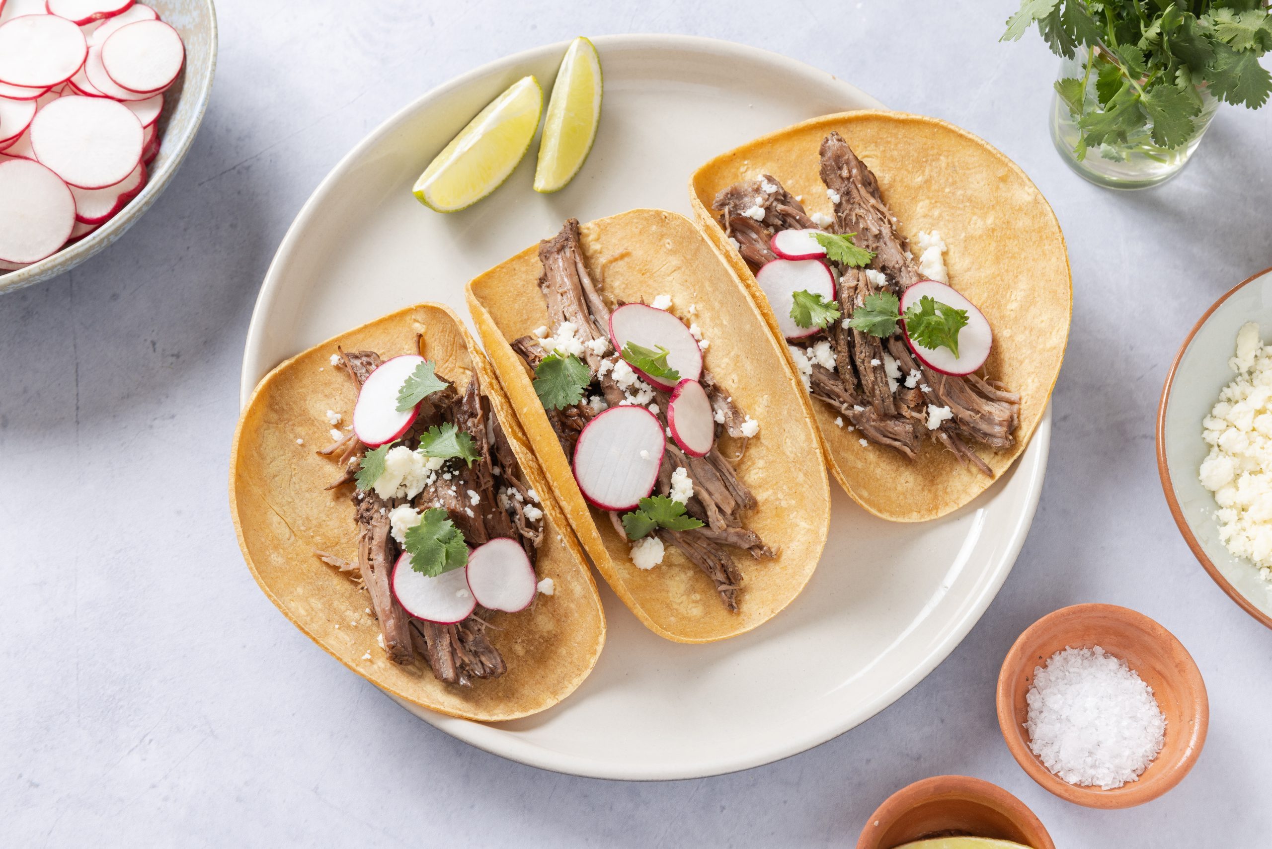 Tacos served on plate with sides of cotija cheese, radishes, lime wedges and cilantro.