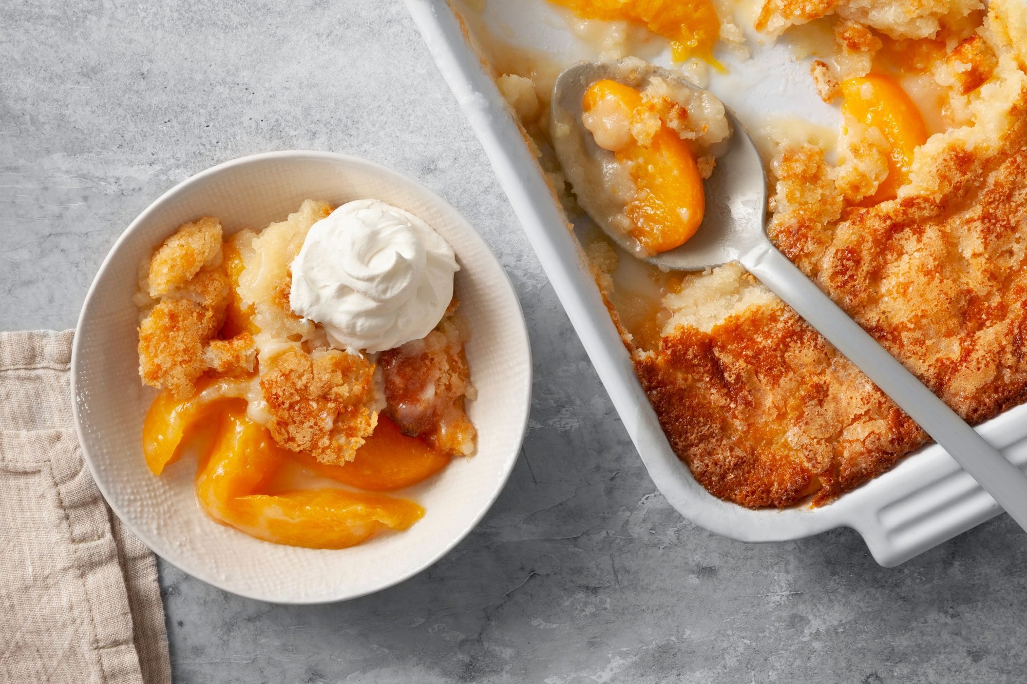Overhead shot of Peach Cobbler; in baking pan; served in a bowl with whipped cream; serving spoon; napkin; grey marble surface;