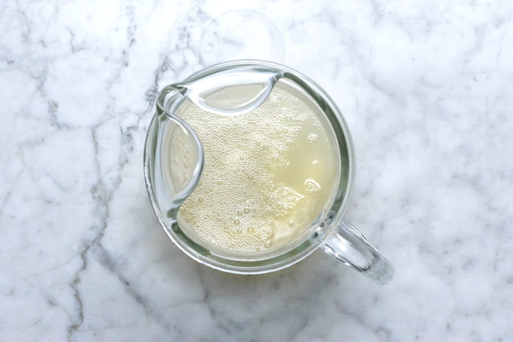 A top-down view of a glass jug filled with frothy, light-colored liquid placed on a white marble surface. 