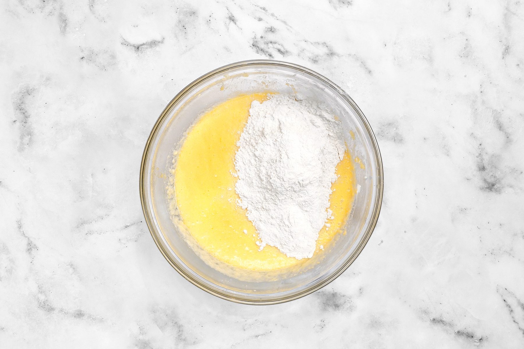 A glass bowl containing a yellow batter mixture with a heap of white flour on top, sitting on a white marble countertop.