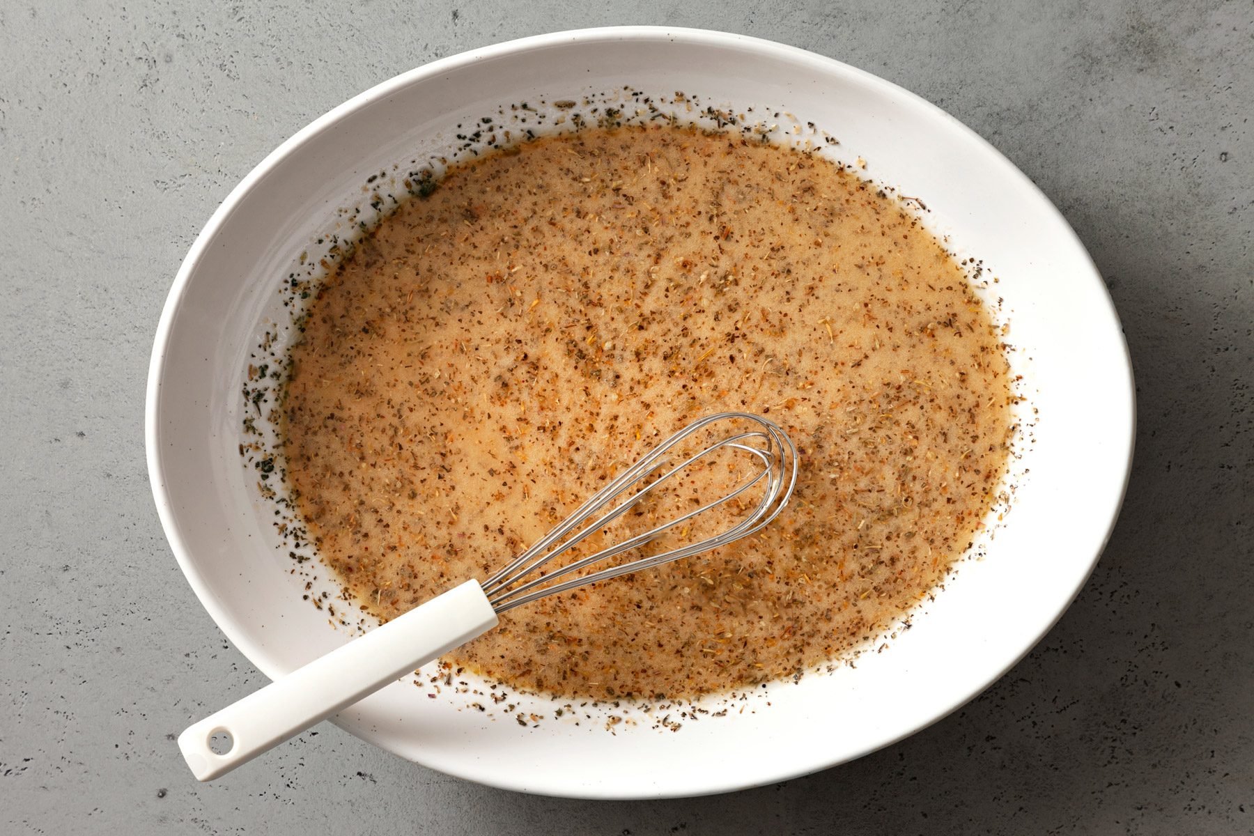 Marinade ingredients mixed in a white bowl against a grey background. A whisker is dipped in the marinade.
