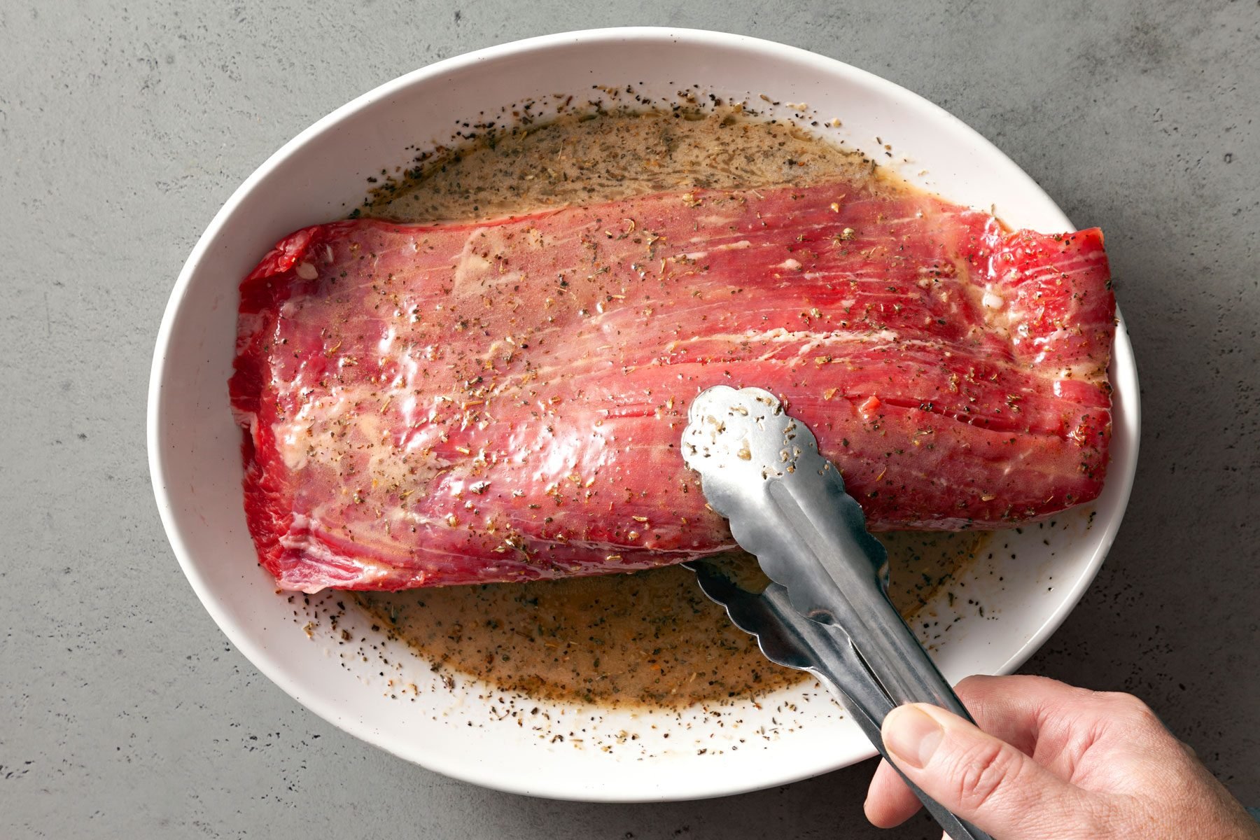 A piece of flank steak is being held by tongs over the marinade in a white bowl against a grey background.