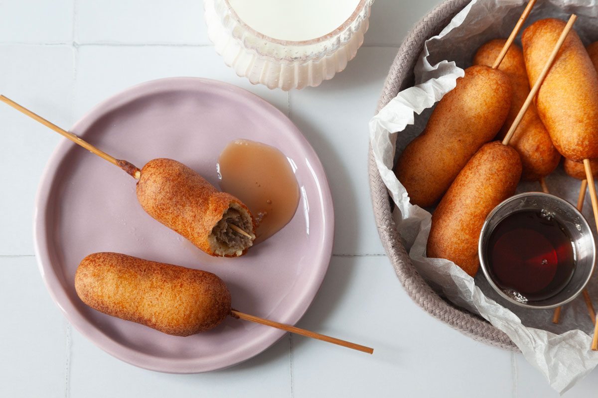 Close up of single serving of Taste of Home Pancakes and Sausage on a Stick on a pink plate with a glass of milk and the full yield peaking in