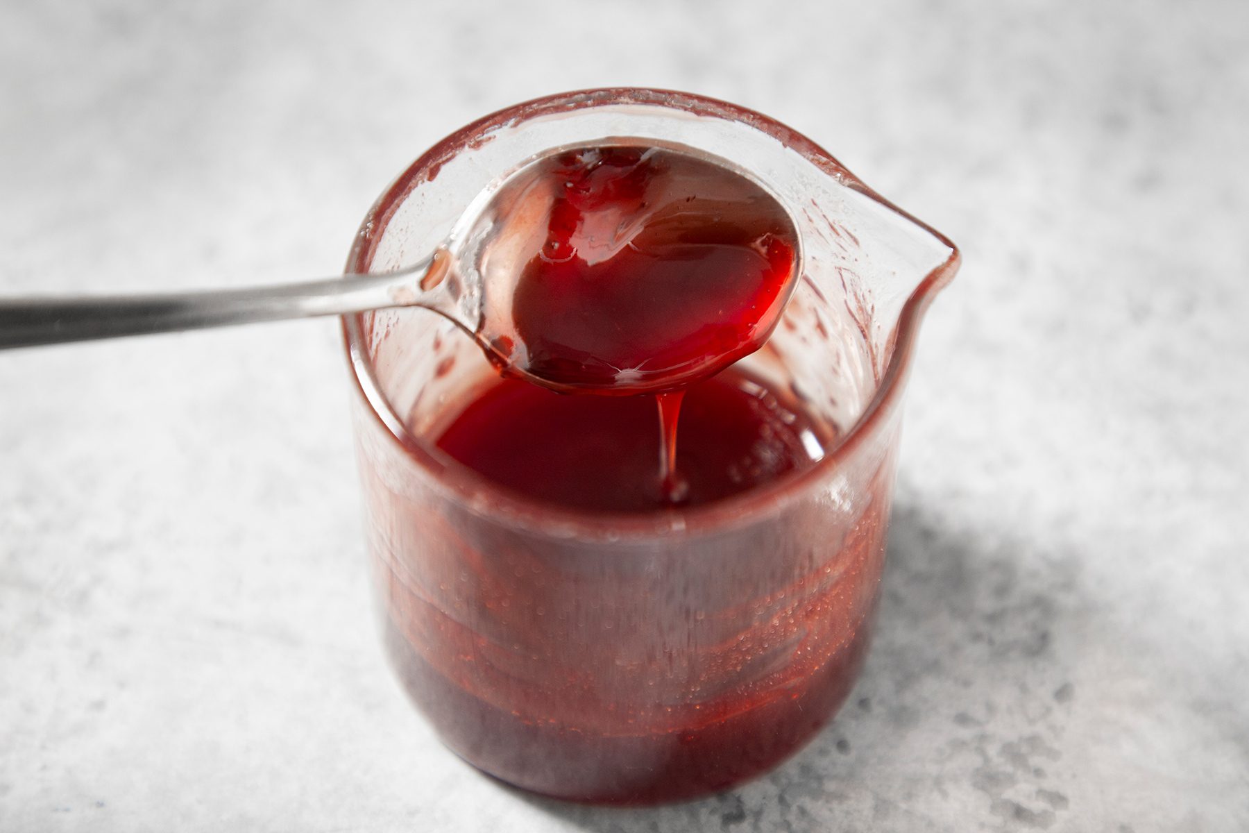 A spoon dipped into a clear glass container filled with red liquid, for serving or measuring. The glass container has a small pour spout. The background is a light, speckled surface.