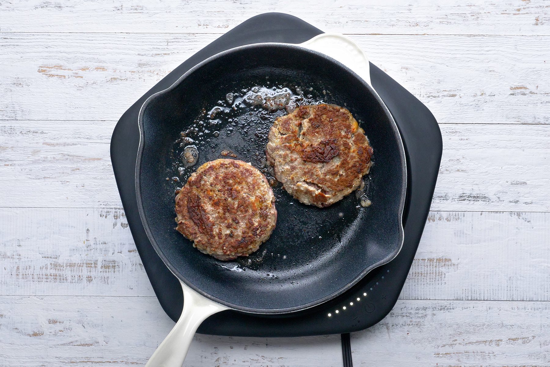 A white skillet with a pair of cooked hamburger patties sits atop a black trivet on a rustic white wooden surface. 