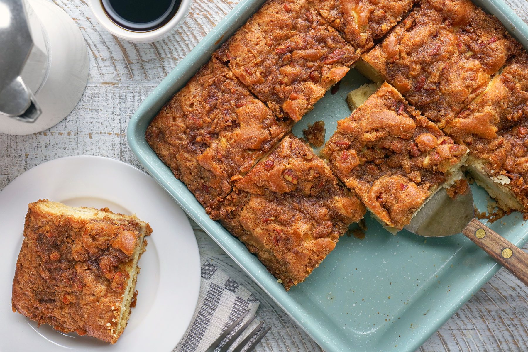 A top view of a baking dish filled with pieces of cake with a serving spatula. One piece has been placed on a white plate next to a cup of coffee, a fork, and a napkin, all on a light wooden surface.