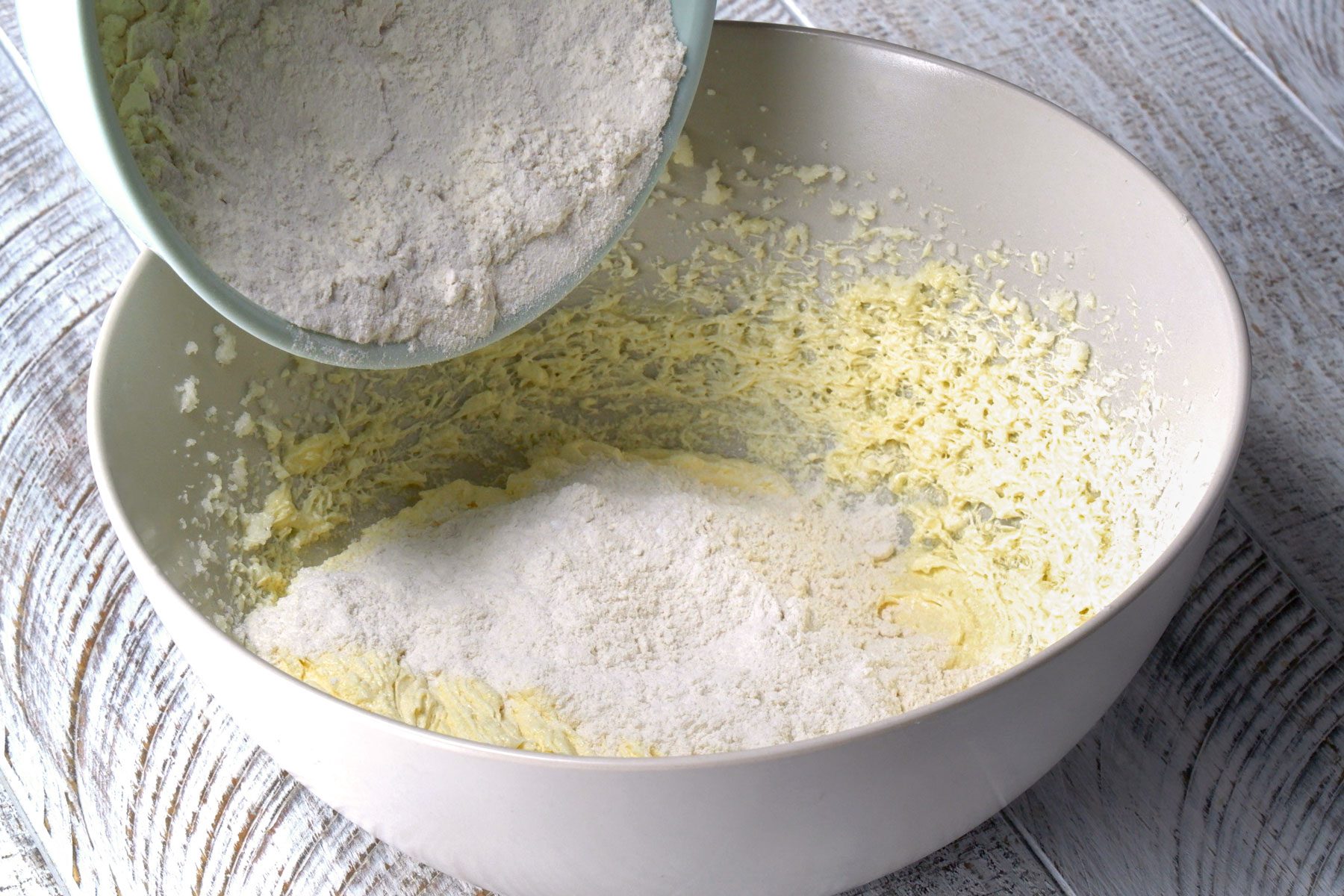 Flour from a small bowl is being added to a big bowl to create a mixture with baking soda and salt.