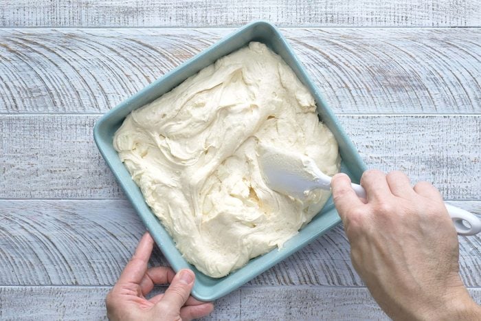 A person's hands can be seen scraping the coffee cake batter into a greased 9-inch square baking pan. The batter is spread evenly to all four corners and edges.