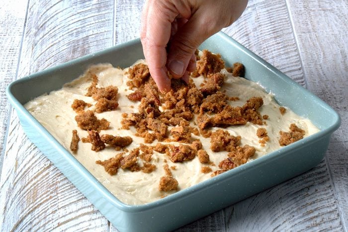 A hand is visible scattering a topping made of brown sugar, pecans, softened butter and apple pie spice evenly all over the cake batter.