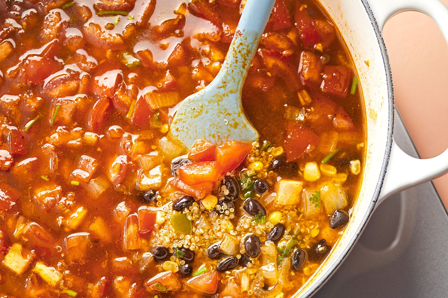A pot of vibrant vegetable stew is being stirred with a blue spoon. The stew contains visible chunks of tomatoes, beans, corn, and other vegetables in a richly colored broth. The image focuses on the hearty, colorful mixture inside the pot.