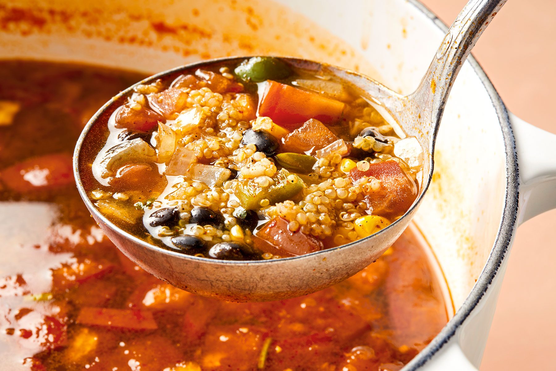 A ladle filled with a hearty soup containing vegetables, beans, and quinoa is being held over a white pot. The soup features various visible ingredients, including diced carrots, green beans, and other vegetables, immersed in a rich broth.