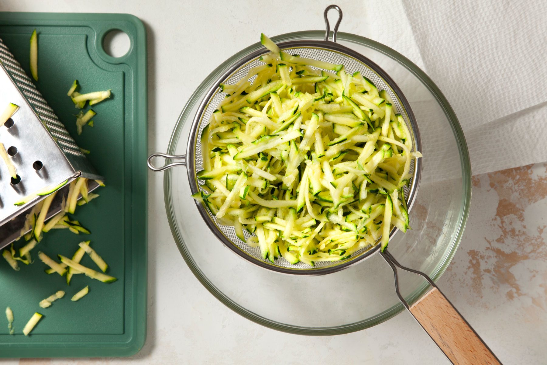 Freshly grated zucchini sits in a mesh strainer over a glass bowl, next to a green cutting board with a box grater on it.