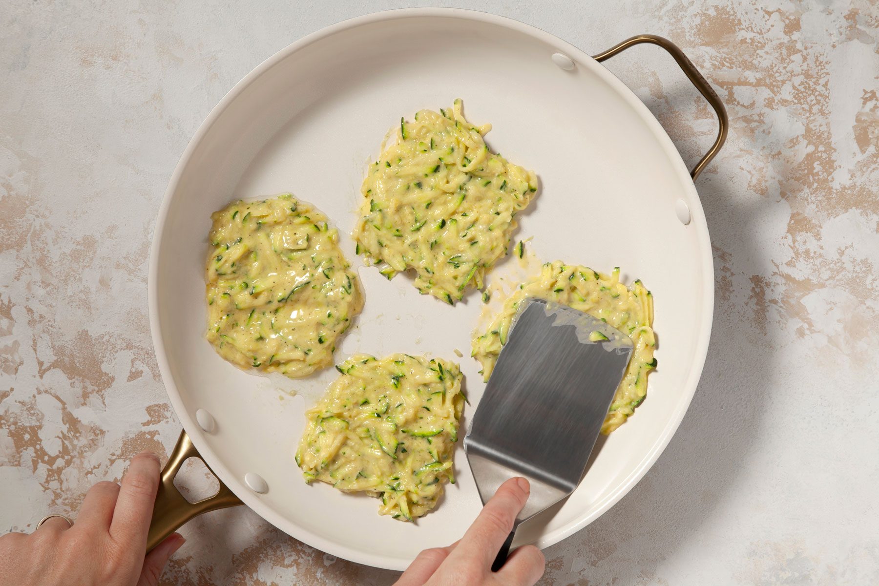A person is cooking four zucchini fritters in a skillet. The person is using a spatula to press one of the fritters. The background surface is light and textured.
