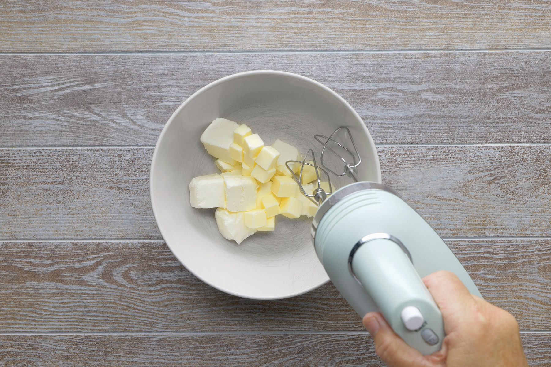 overhead shot; grey background; A white bowl contains a mixture of butter and sugar; An electric mixer is being used to beat the mixture together; The butter is cut into cubes and the mixture is a pale yellow color