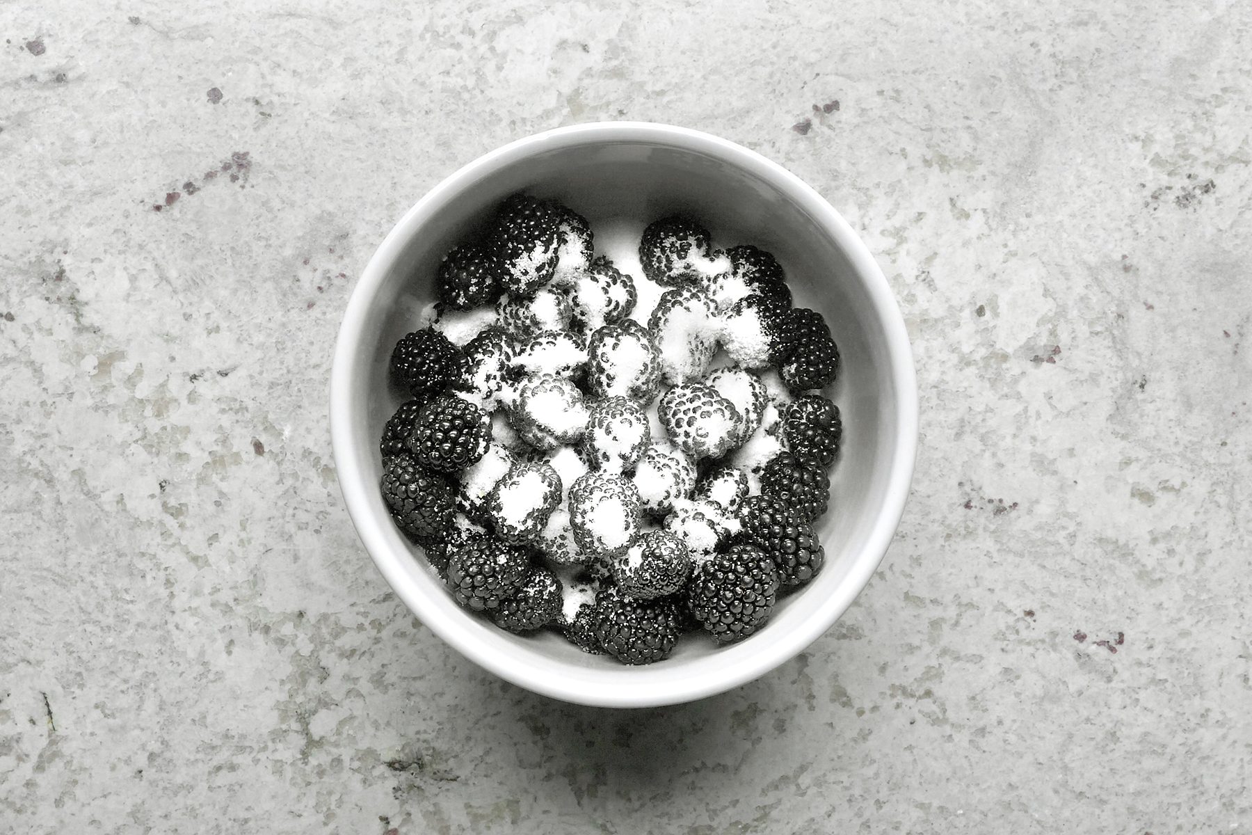 A white bowl filled with blackberries lightly dusted with powdered sugar sits on a textured, light gray surface. The contrast of dark berries and white sugar creates a visually striking effect.
