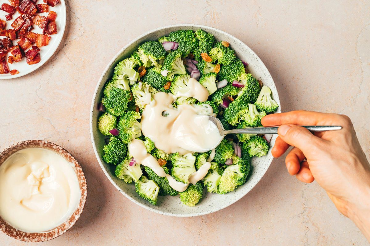 Hand pouring Miracle Whip dressing over Broccoli Raisin Salad Recipe for Taste of Home