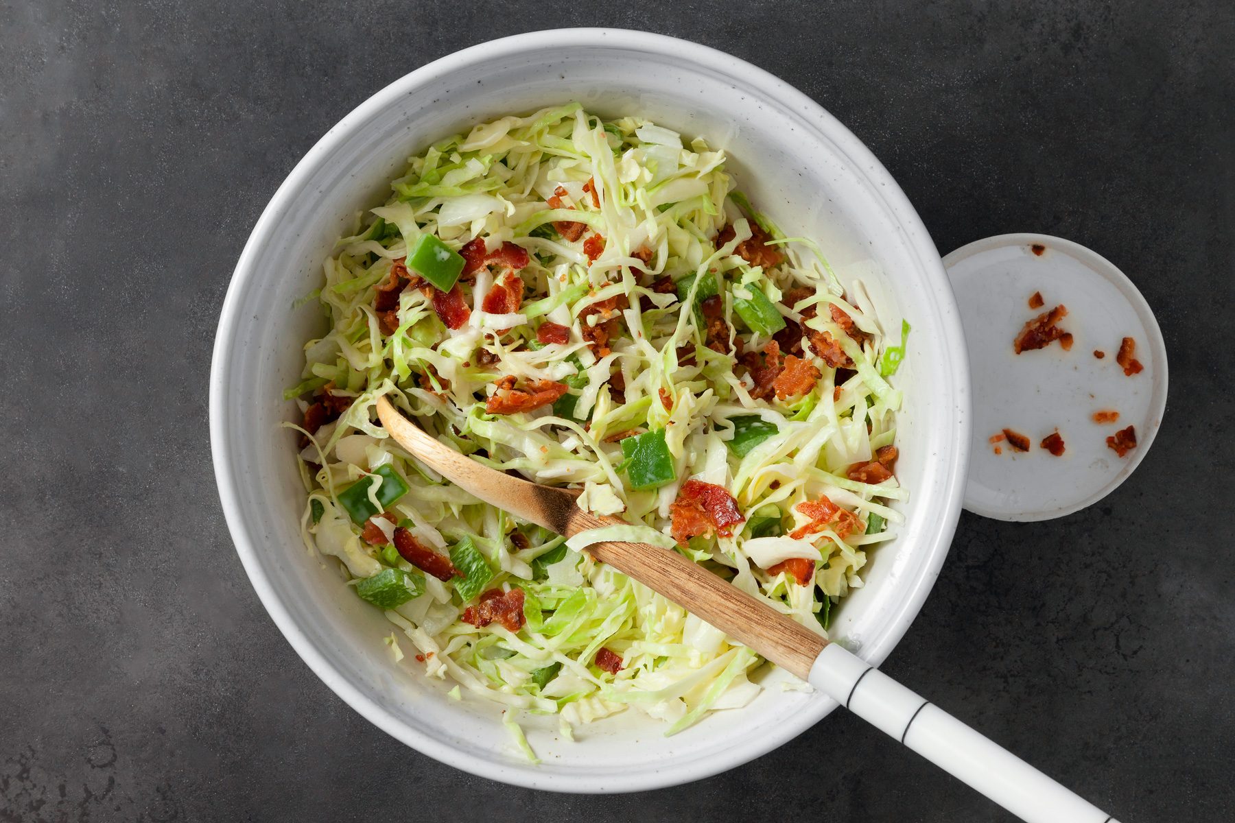 overhead shot; dark grey background; prepared Favorite Cabbage Salad in the white bowl with some sprinkled chilli flakes and a wooden spatula is placed inside the bowl